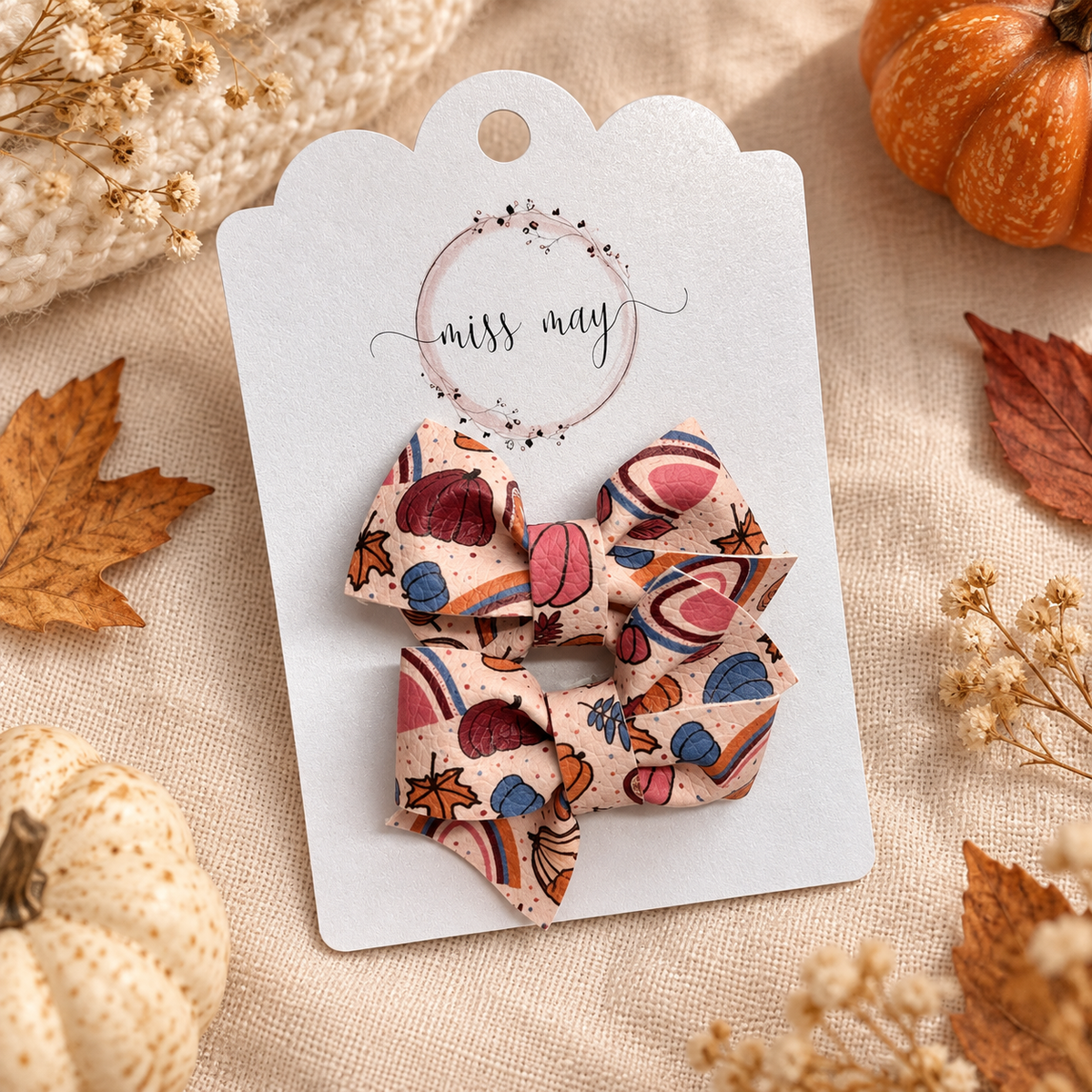 Two pumpkin floral rainbow pigtail bows on a Miss May display card, arranged on cream linen with pumpkins, dried flowers, and autumn leaves.